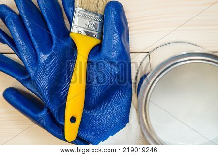 Protection for redecoration work. A pair of blue latex gloves, a brush and a tin can of white oil paint on a light uncolored wooden background. Close up. Top view. Space for your text or pruduct display.
