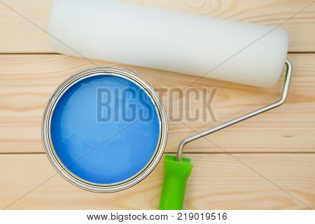 A can of blue oil paint and a clean paint roller on a light uncolored wooden background. Close up. Top view. Space for your text or pruduct display.