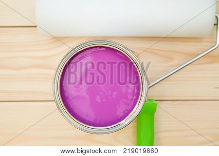 A can of fuchsia oil paint and a clean paint roller on a light uncolored wooden background. Close up. Top view. Space for your text or pruduct display.