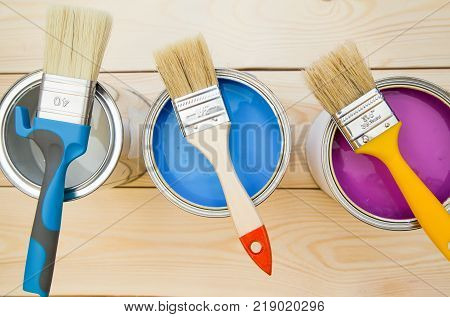 Ready to paint a house. Three tin cans of household paint in a row with brushes on top, light uncolored wooden background. Close up. Top view. Space for your text or pruduct display.