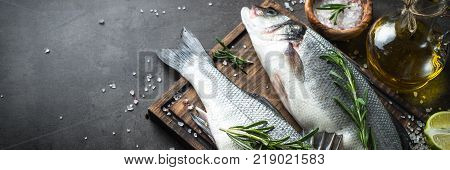 Fresh fish seabass and ingredients for cooking. Raw fish seabass with spices and herbs on black slate table. Long banner format.