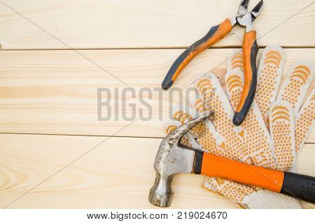 Tools for house repair- a hammer, pilers and a pair of protective work gloves on a light uncolored wooden background. Top view. Space for your text or pruduct display.