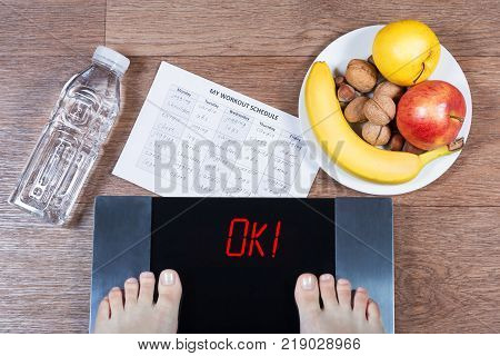 Female feet on digital scales with word ok surrounded by bottle of water, plate with healthy food and workout schedule paper. Concept of active and healthy lifestyle. Top view.