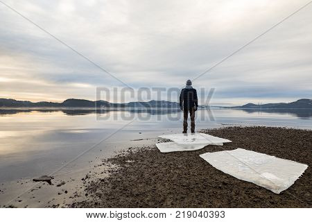 Man standing at the beach in the Norwegian fjord Tovdalsfjorden. Ice floes on dark sand, calm sea, mist and fog. Hamresanden, Kristiansand, Norway
