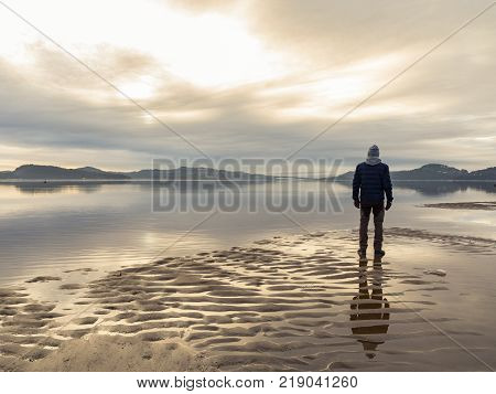 Man standing at the beach in the Norwegian fjord Tovdalsfjorden, looking at the calm sea and the mist and fog. Sand patterns and reflections of the man in the water. Hamresanden, Kristiansand, Norway