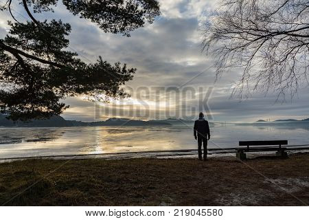 Man standing next to a bench at the beach in the Norwegian fjord Tovdalsfjorden, looking at the calm sea and the mist and fog. Hamresanden, Kristiansand, Norway