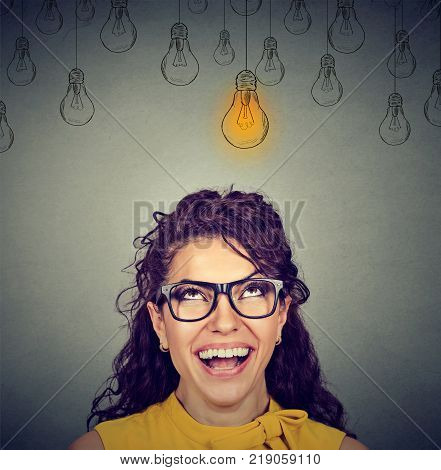 Portrait happy beautiful woman in glasses looking up at idea light bulb above head isolated on gray wall background
