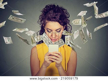 Online banking money transfer e-commerce concept. Happy young woman using smartphone with dollar bills flying away from screen isolated on gray wall background.