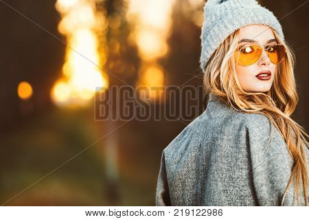 Autumn fashion. Fashionable young woman wearing a coat and a woolen hat posing in the park at a sunset.