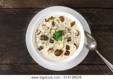 An overhead photo of a plate of a mushroom cream soup with champignons, cremini, fresh parsley, with grated cheese, on a dark rustic background texture, with a place for text