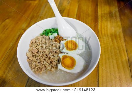 Minced pork congee with egg fresh ginger and coriander on white cup and spoon over the wood table. Second view.