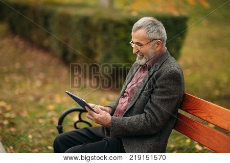 Grandpa use a tablet sitting in the pakr on the bench.
