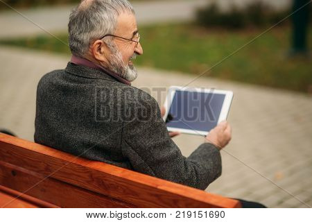 Grandpa use a tablet sitting in the pakr on the bench.