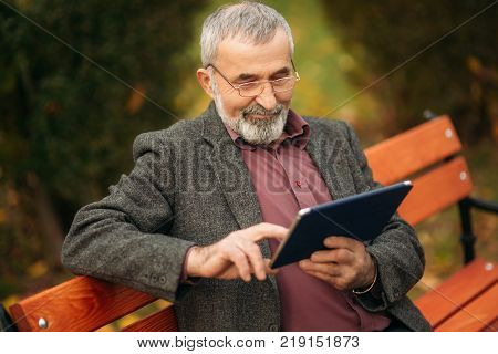 Grandpa use a tablet sitting in the pakr on the bench.