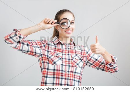 smiling woman in a plaid shirt looks through a magnifying glass. gesture thumbs up