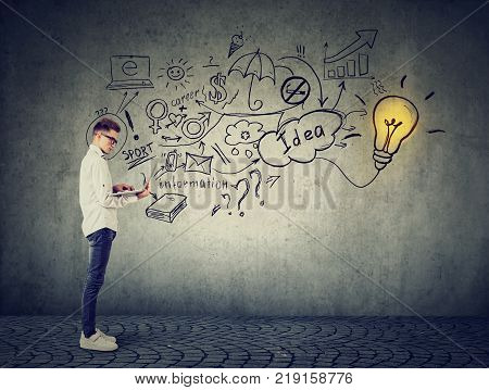 Young man using a laptop with light bulb plugged in it and info graphic plan written on concrete wall