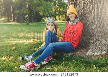 Outdoor portrait of beautiful female dressed in warm cap and warm loose sweater spend weekends together with her little daughter sit on green grass near tree enjoy fresh air and wonderful nature