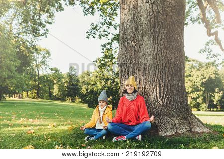 Yoga and meditation concept. Beautiful young woman in knitted clothes and small child keep eyes closed meditate outdoors in green park feel relaxed calm and peaceful keep hands in mudra sign