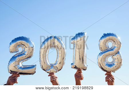 men hands holding some silvery number-shaped balloons forming the number 2018, as the new year, against the blue sky with a blank space on top