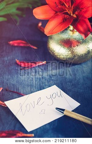 I Love You written on a peace of paperr pen and golden vase with red lily and petals on the dark wooden background with retro filter effect. Selective focus. space fpr text