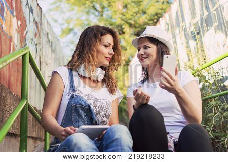 two adult girls sitting in the shade on the stairs listening to music and photographing with a tablet