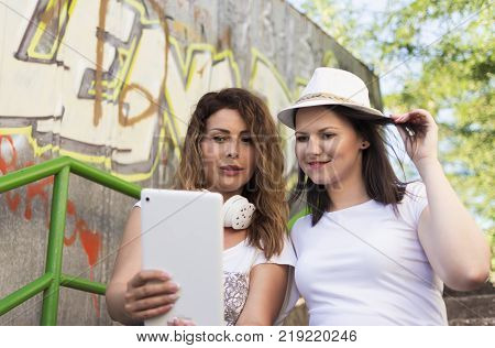 two adult  girls sitting in the shade on the stairs, listening to music and photographing with a tablet