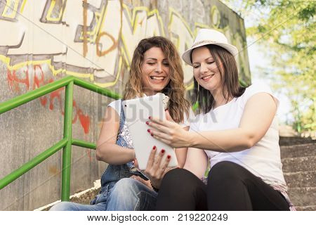 two adult girls sitting in the shade on the stairs listening to music and photographing with a tablet