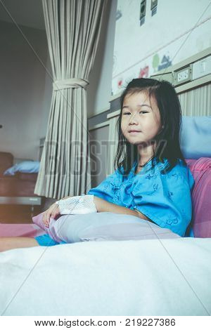 Illness asian child admitted in hospital while saline intravenous (IV) on hand. Unhappy girl feeling sad bored kid emotion. Health care stories. Vintage film filter effect.