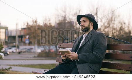 Creative young writer man in hat and coat write notes for his future book with pen in notebook sitting on city street bench