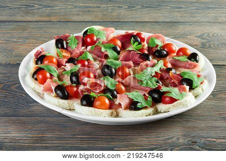 Delicious restaurant appetizer : canapees with white bread, bacon rolls, fresh cherry tomatoes and green parsley leaves. Tasty dish for catering or buffet. A plate stands on the wooden table