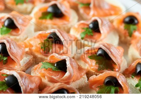Little canapees with smoked salmon rolls and black olives on the white bread, covered with butter and decorated with fresh green parsley leaves.