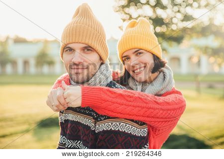 Smiling glad young female in hat and warm cotton sweater embraces her husband who stands back enjoy spending weekends together have walk in garden or park admire splendid sunny autumn weather