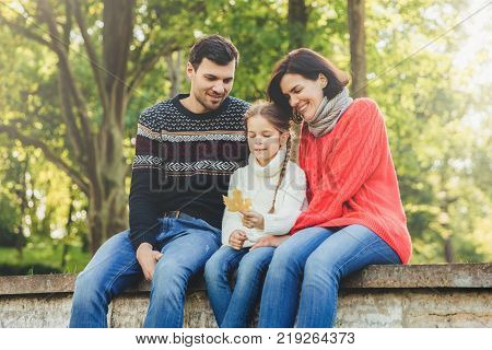 Adorable cute lovely small chld sits between her parents outdoors holds leaf in hands spend weekends together. Friendly young family enjoy togetherness have happy expressions sit on bridge