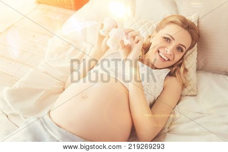Long lasting anticipation. Cheerful delighted pregnant woman lying in bed and holding clothes for baby while resting at home