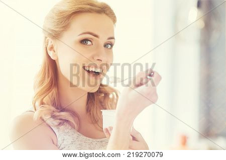 Yammy yammy. Cheerful content beautiful woman holding spoon with joghurt and eating it while feeling happy