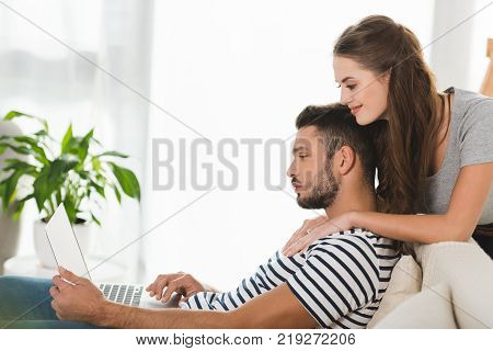 side view of young woman embracing her boyfriend while he working with laptop at home