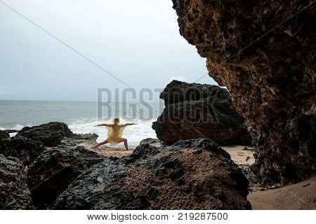 Woman in rain coat doing stand yoga asana on ocean beach during storm. Concept of yoga travelling on Bali