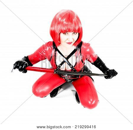 Attractive fetish cosplay woman holding a whip. Wearing red and black latex rubber catsuit and red wig sitting on haunches. Filters applied for artistic effect. White background.