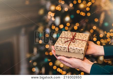 Hirizontal shot of beautiful wrapped present box in woman`s hands against decorated Christmas or New Year tree. New Year background. Focus on gift box. Celebration concept.
