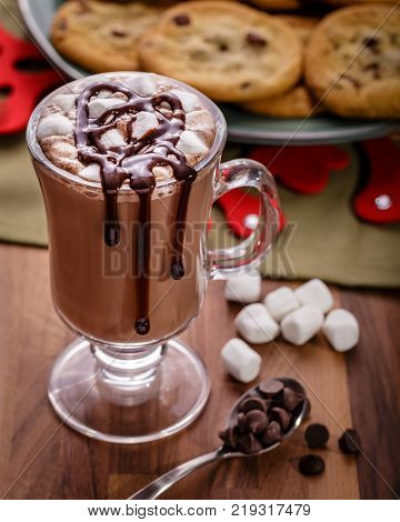 Frech Hot Chocolate with a handful of mini marshmallows and chocolate syrup drizzled on top. A plate of homemade chocolate chip cookies is in the back ground