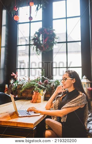 Beautiful Young Brunette Woman Inside Cafe With Christmas Interior At Wooden Table By The Window Tal