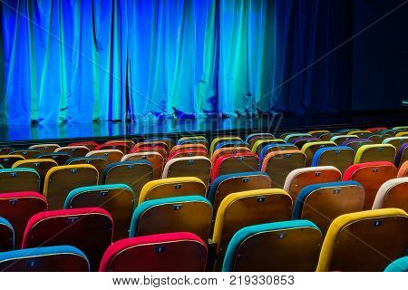 The auditorium in the theater. Blue-green curtain on the stage. Multicolored spectator chairs. Lighting equipment.