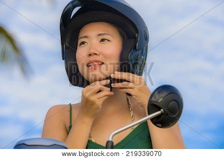 young pretty happy and cute Asian Chinese woman adjusting motorcycle helmet riding on scooter motorbike isolated on a blue sky in safe bike ride safety and security driving concept