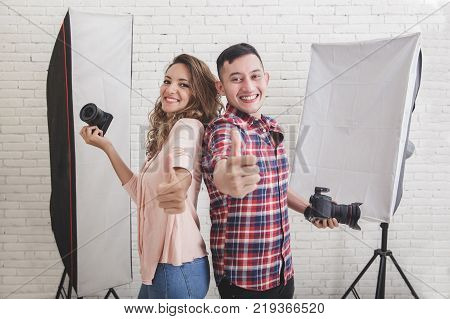 portrait of two young photogaphers giving thumbs up in studio with lighting equipped at the background