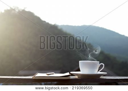 cup of hot coffee for brackfast and note book with pen on wooden balcony near river and mountain background