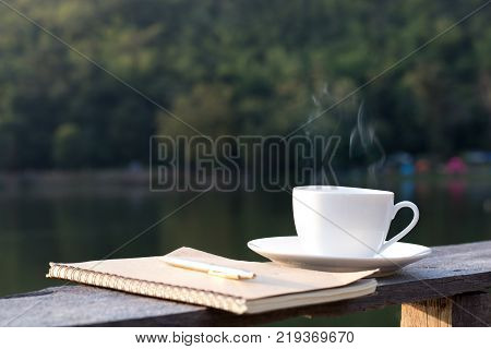 cup of hot coffee for brackfast and note book with pen on wooden balcony near river and mountain background
