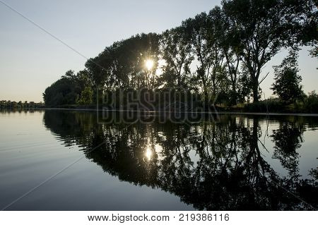 Trees on the shore are reflected on the surface. The sun is making its way to the lake through the branches. Cod and dry branches the right border the coast and water.