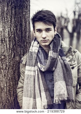 Handsome young man outdoor in winter fashion, wearing dark jacket and woolen scarf in city park looking at camera