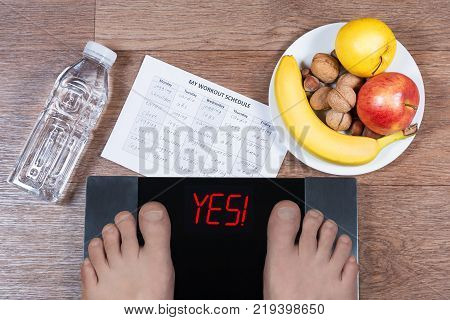 Male Feet On Digital Scales With Word Yes Surrounded By Bottle Of Water, Plate With Healthy Food And