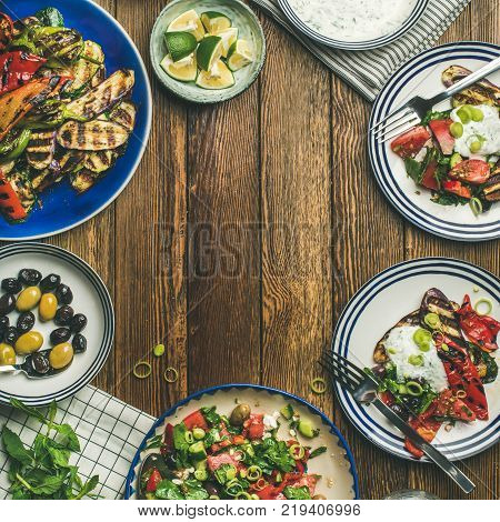 Flat-lay of healthy dinner table setting. Fresh salad, grilled vegetables with yogurt sauce, pickled olives over wooden background, copy space, selective focus, square crop. Clean eating, vegan food